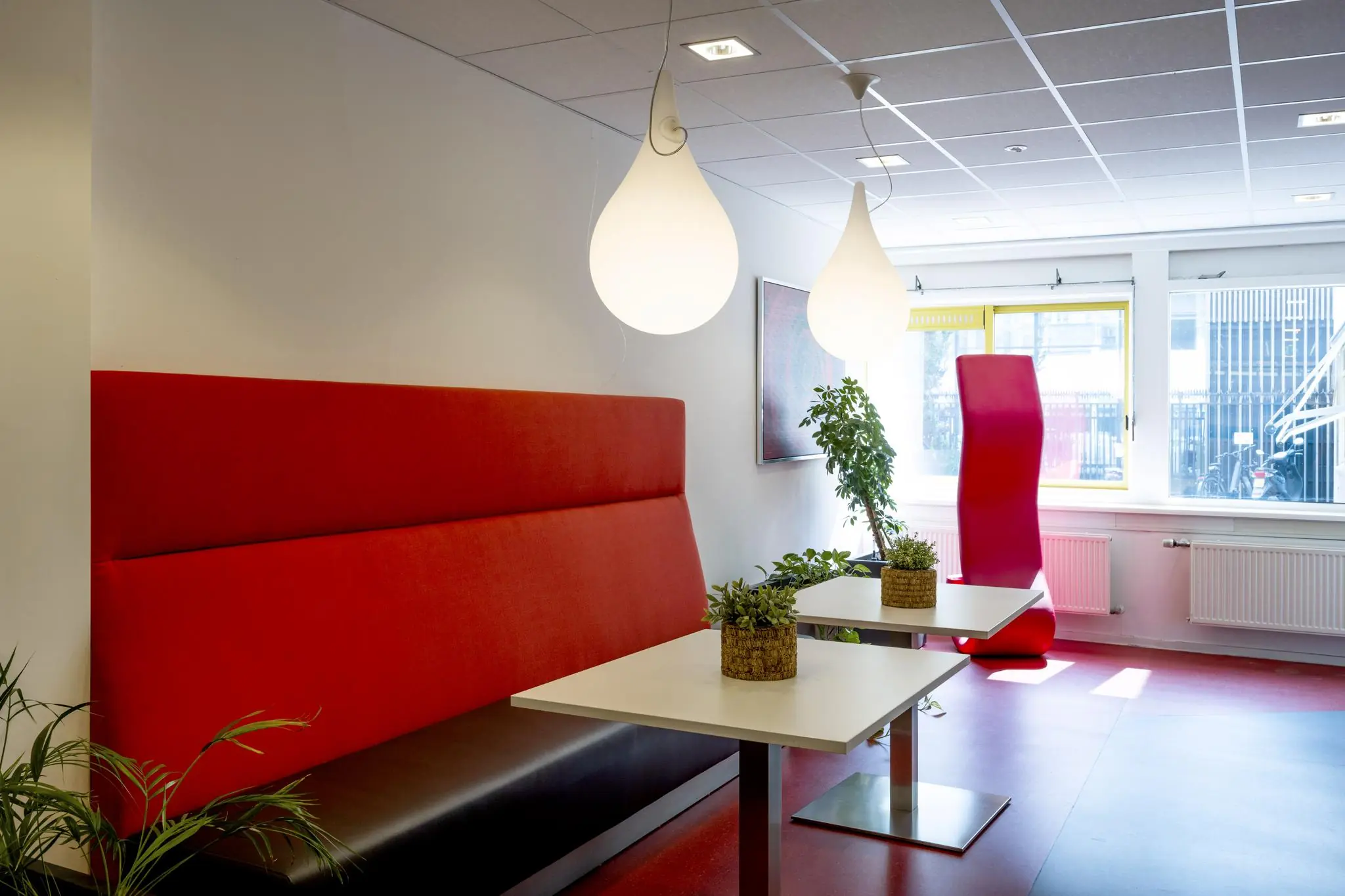 Moderne wachtruimte aan de Jan Tooropstraat met rode zitbanken, witte tafels, hanglampen en planten.